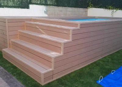 Piscina elevada forrada con madera tecnológica clara, con escalera ancha y puerta de registro, en un jardín de Madrid