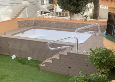 Piscina elevada recién forrada con madera tecnológica, con escalera, puertas de registro y barandillas de acero, en un jardín de Murcia
