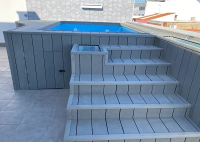 Piscina elevada forrada con lamas verticales de madera tecnológica gris, con escalera y puertas de registro, en Badajoz