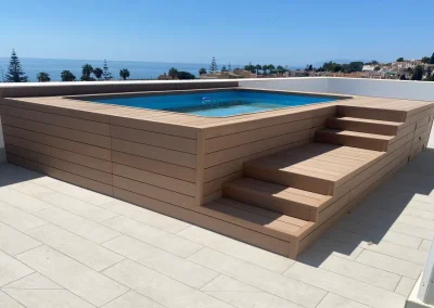 Piscina elevada forrada con madera tecnológica clara y escalera integrada, en una terraza con vistas al mar en Málaga