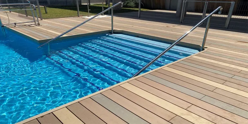 Tarima de madera tecnológica en varios tonos alrededor de una piscina, con escalera de obra y barandillas de acero